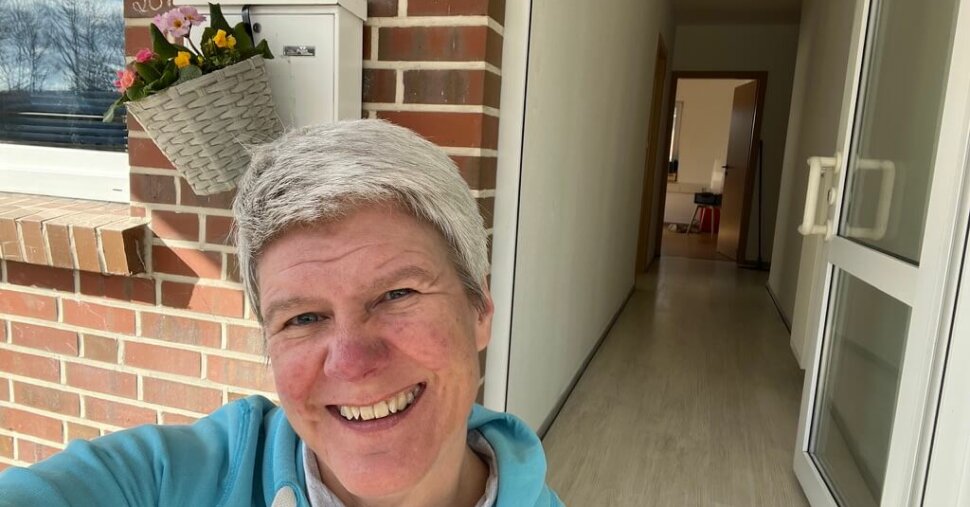 Das Foto zeigt eine lachende Frau und im Hintergrund kann man in einen langen Flur reinschauen.