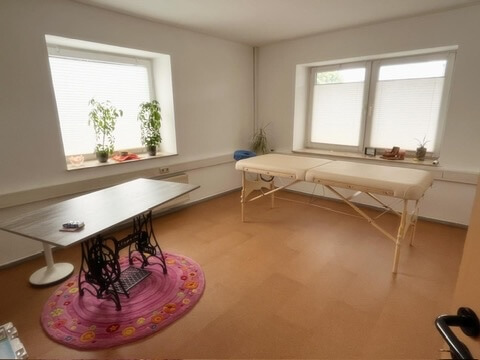 Das Bild zeigt einen Raum mit einem Schreibtisch und einer Massageliege darin. Plissees sind an den 2 Fenstern.