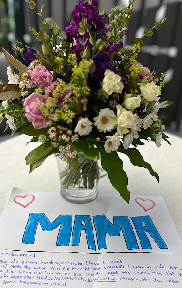 Das Bild zeigt einen bunten Blumenstrauß und davor ein gemaltes Bild mit dem Schriftzug „Mama“ und die Beschreibung dazu.