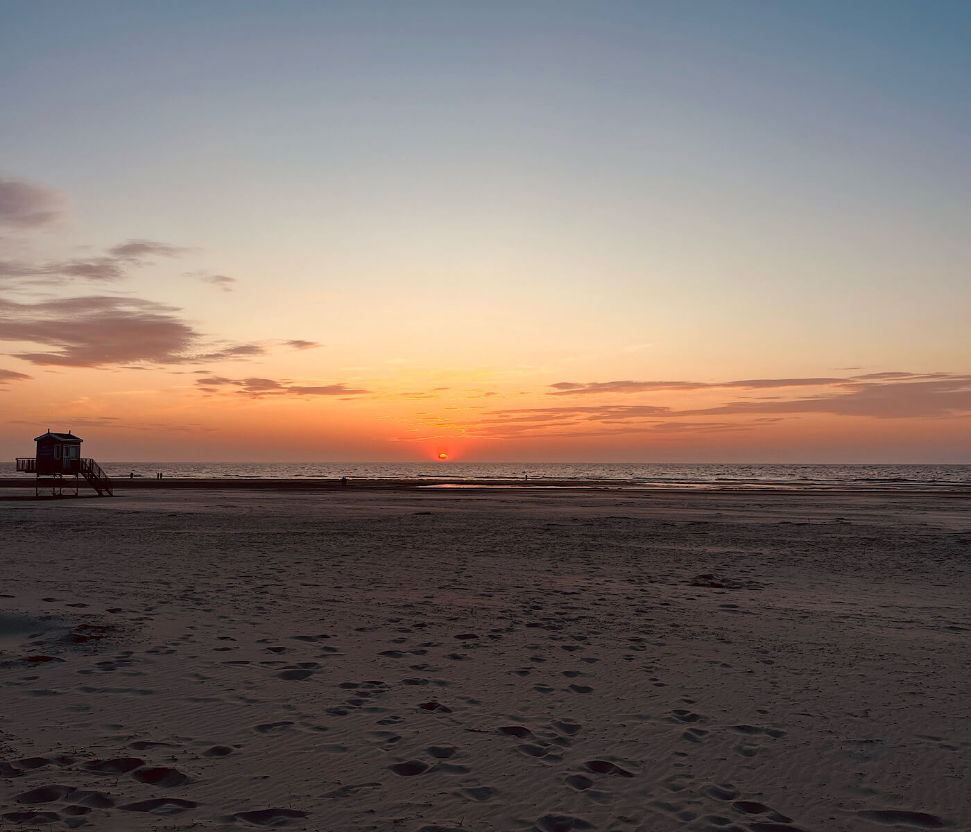 Das Bild zeigt einen Sonnenuntergang am Strand. Ein DLRG Turm steht im hintergrund links.